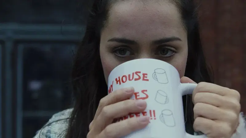 Marta Cabrera stands on the balcony holding Harlan's "My House, My Rules, My Coffee!!" mug